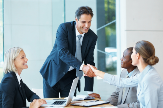 Business meeting in a bright office: a man in a suit stands and smiles while shaking hands with colleagues around a conference table with laptops and documents.