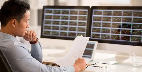 Man in business attire analyzes market data across multiple monitors, holding data sheets, illustrating the rise of marketing analytics in finance.