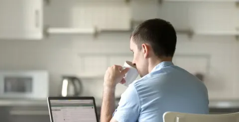Man in a light blue shirt sits at a kitchen table with a laptop, drinking from a cup, suggesting remote learning or online study in a home environment.