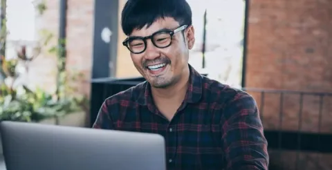 Man in glasses and a plaid shirt works on a laptop in a casual, modern work environment, conveying real-world financial application.