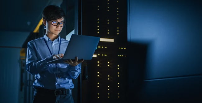 IT professional checking a laptop in a data center with blue lighting and server towers.