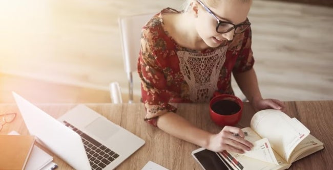Woman in a floral dress sits at a desk with a planner and coffee, using a laptop, suggesting balancing work tasks and study in a home or casual workspace