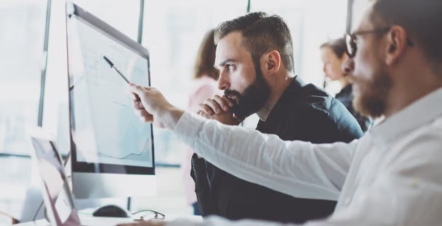 A person is pointing at data on a computer screen while another person attentively observes, suggesting a collaborative work or analysis session.