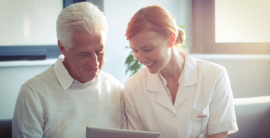 An elderly person and a healthcare professional sit closely together, looking at a laptop in a bright, comforting setting.