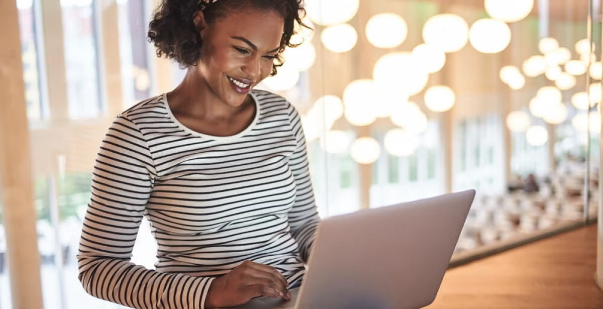 Woman in a striped shirt using a laptop in a brightly lit room, suggesting online research or studying financial aid options.
