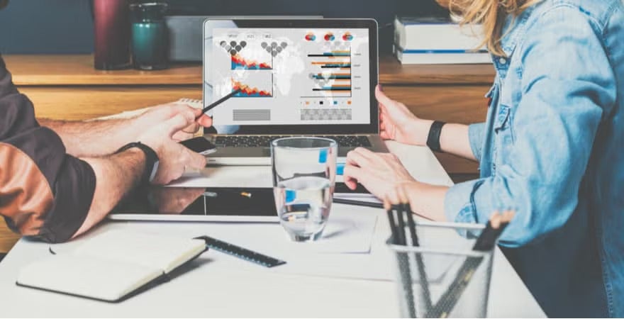 Two people collaborate at a desk, analyzing data visualizations and charts displayed on a laptop screen.