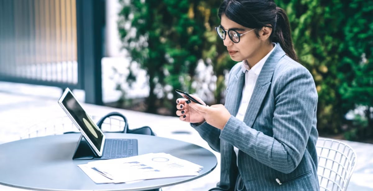 Businesswoman in a pinstripe suit sits outside at a table with a tablet, reviewing financial documents and typing on her phone.